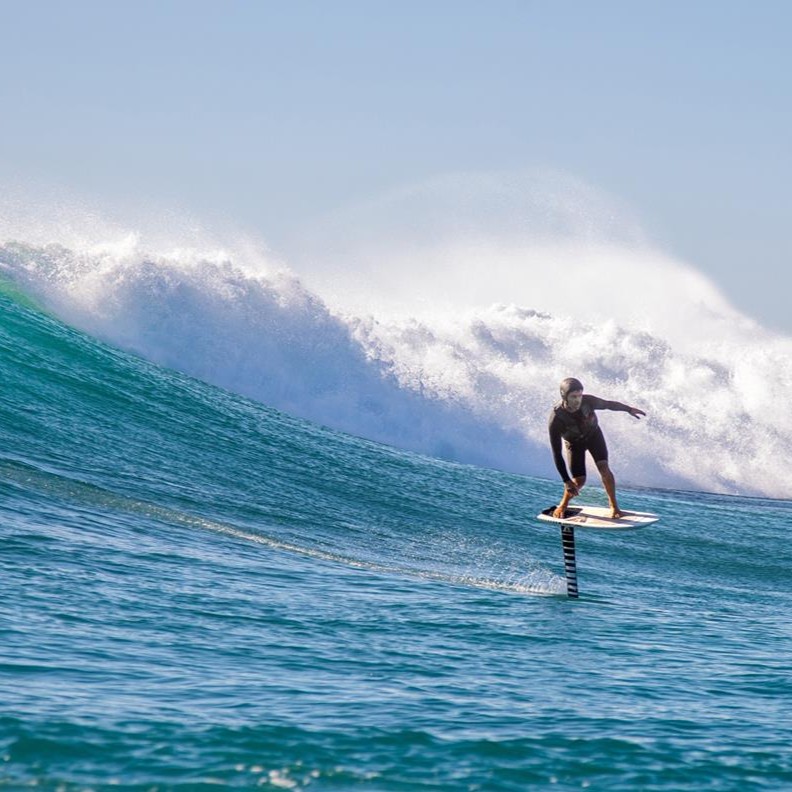 Surfeur en combinaison noire pratiquant le surf foil, sur une planche équipée d'un foil ARMSTRONG Foils - Performance Mast, avec une grande vague en arrière-plan